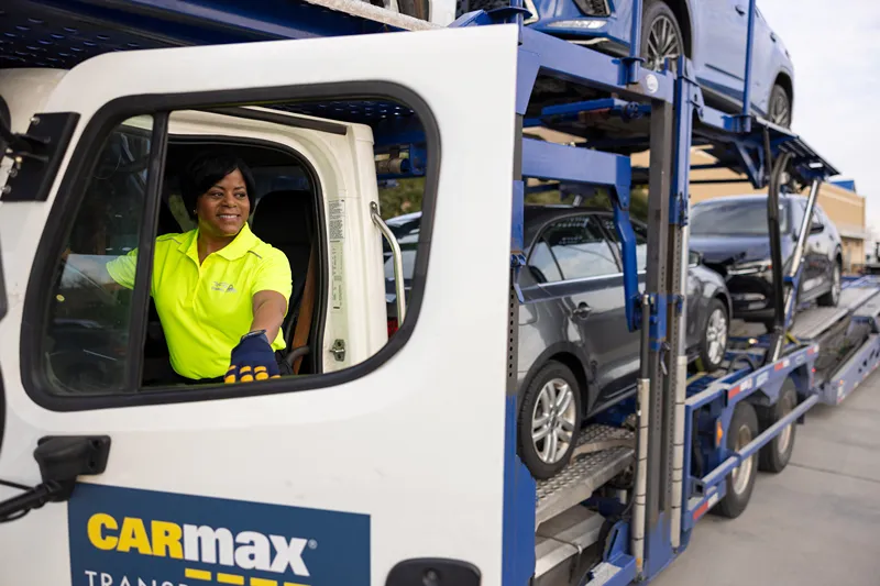 A smiling CarMax truck driver is sitting in the cab of a car transport carrier.