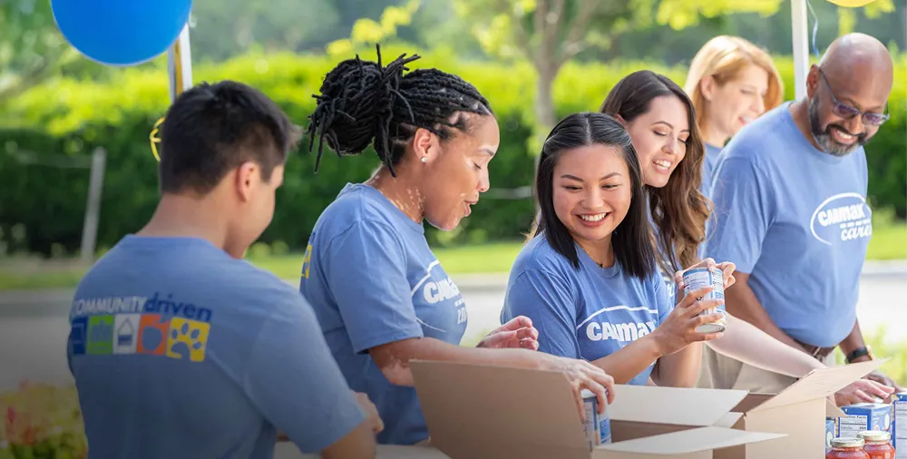 A diverse group of smiling CarMax employees is volunteering by packing food boxes outdoors.