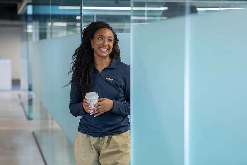 A smiling female student or intern is walking with a cup of coffee in a bright office hallway.