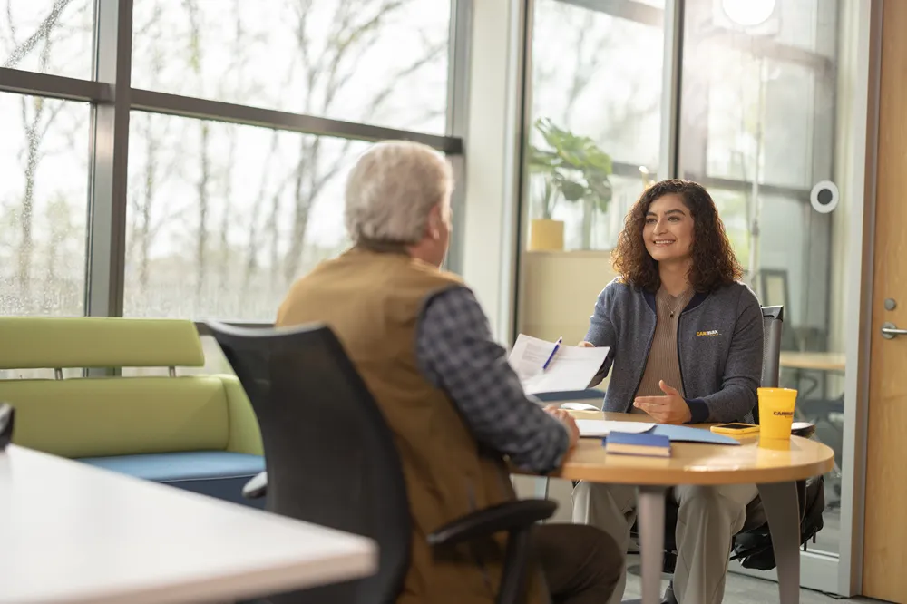 A smiling CarMax finance employee is assisting a customer with paperwork at a table in a modern office.