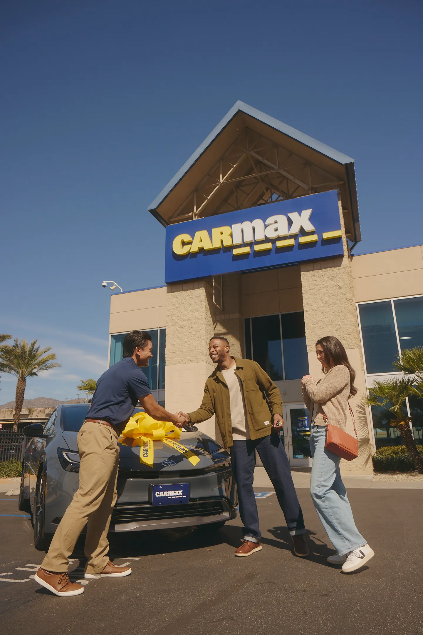 A CarMax employee is happily shaking hands with a customer receiving a new car outside the store.