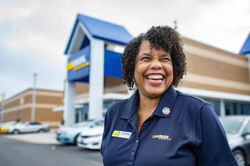 A cheerful CarMax employee with a military affiliation pin is smiling outside a store.