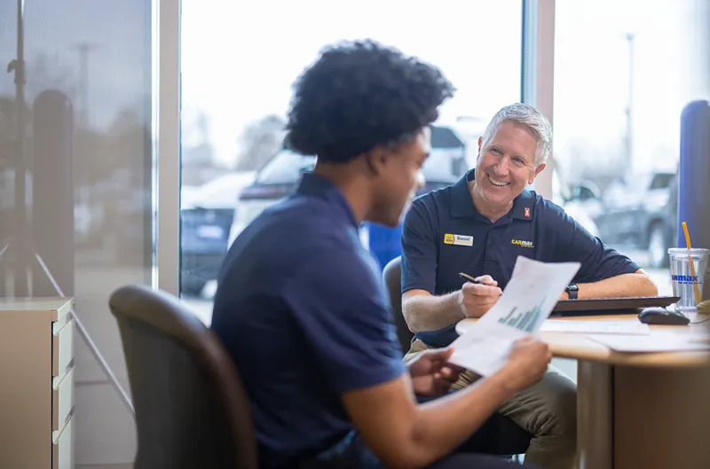 A smiling CarMax mentor is discussing documents with a young colleague in an office.