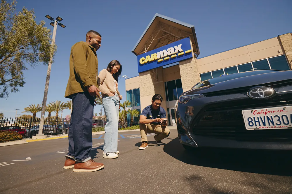 A CarMax employee is inspecting a car for a smiling couple outside a CarMax store.
