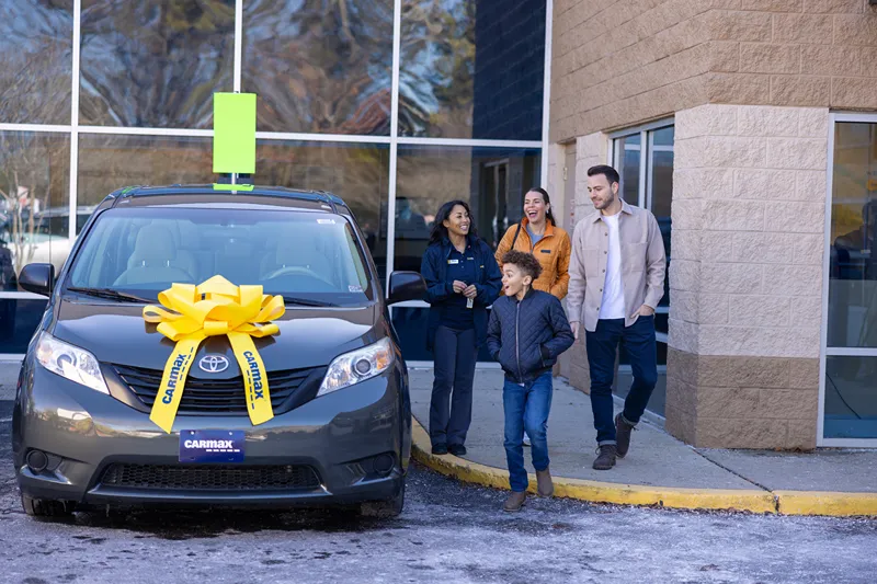 A smiling family is receiving their new car, adorned with a large yellow bow, from a CarMax employee.