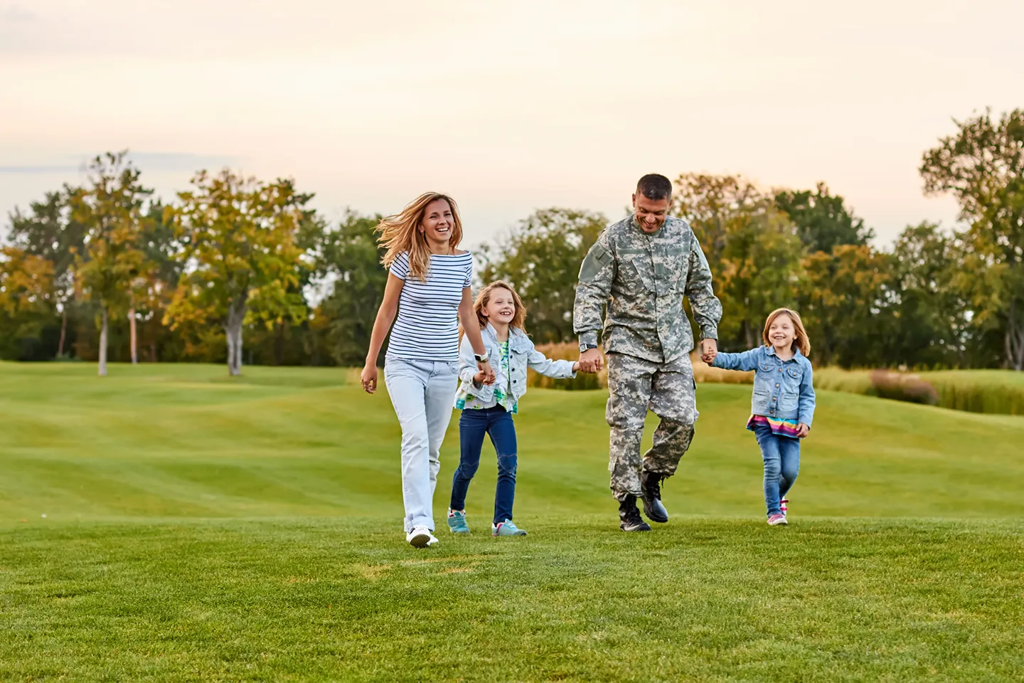 A military service member and his smiling family are walking across a grassy field at sunset.
