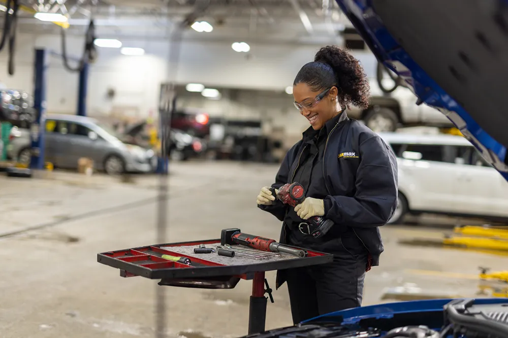 A smiling female auto technician is preparing tools on a cart in a busy service bay.