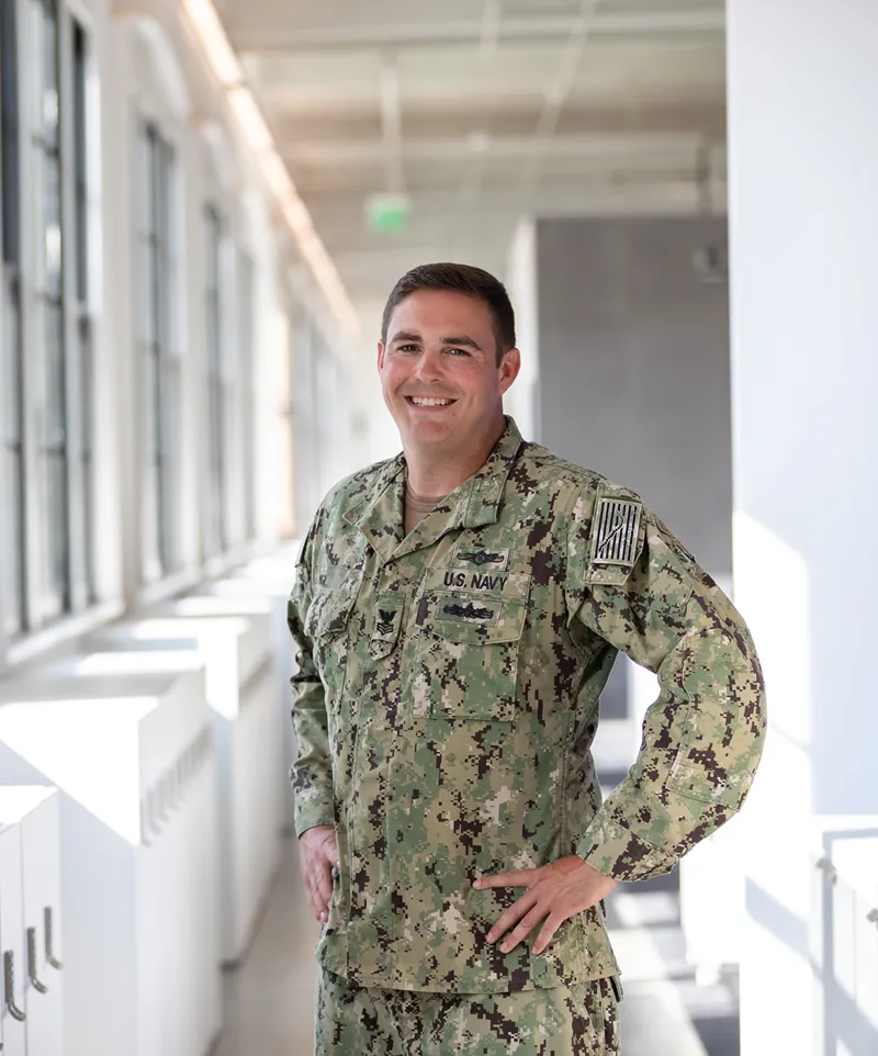 A smiling military veteran in US Navy uniform is standing in a brightly lit office hallway.