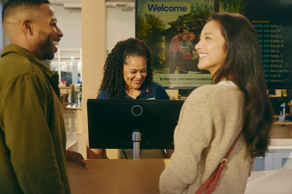 A smiling customer service representative is greeting a happy couple at a service counter.