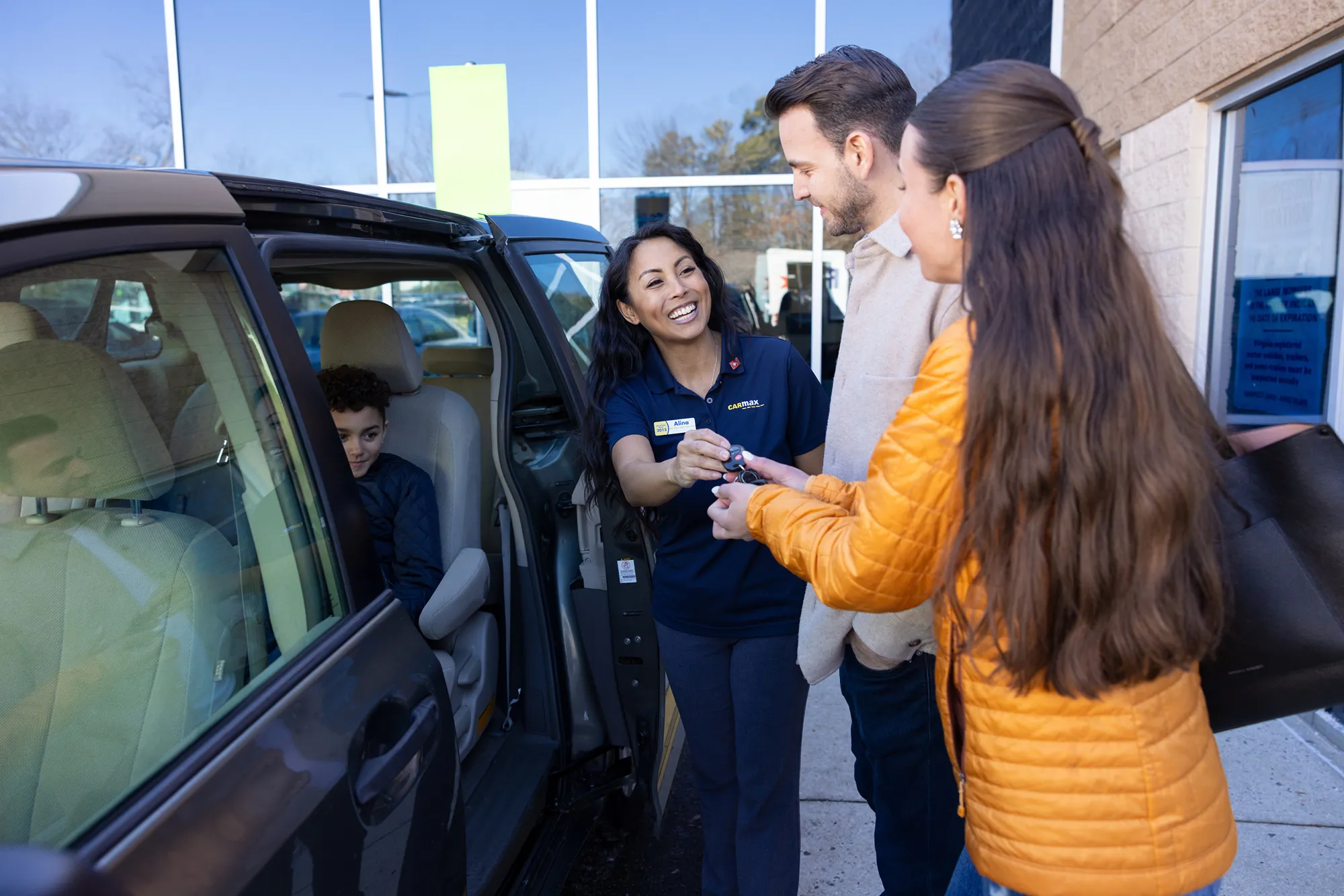 CarMax employee handing car keys to smiling customers outside the dealership, with a child sitting in the back seat.