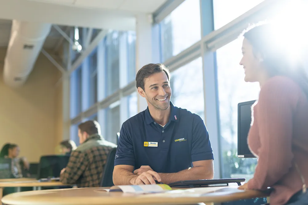 A smiling CarMax employee is happily assisting a customer at a desk near a window.
