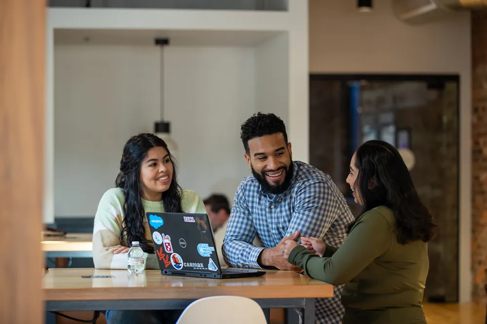 Three smiling CarMax technology professionals are collaborating around a laptop