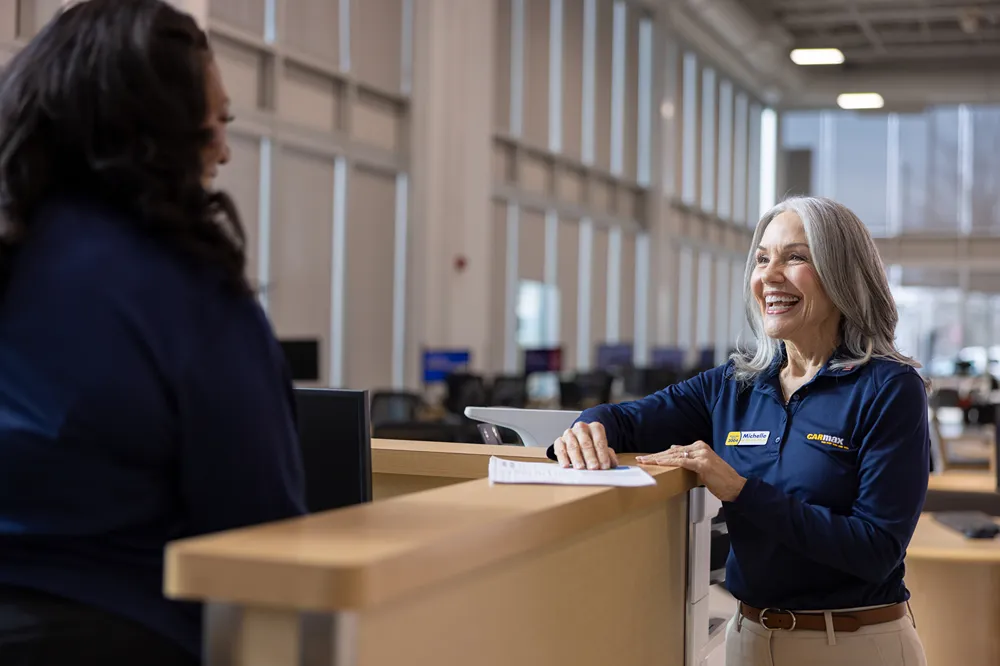 CarMax employee handing car keys to smiling customers outside the dealership, with a child sitting in the back seat.