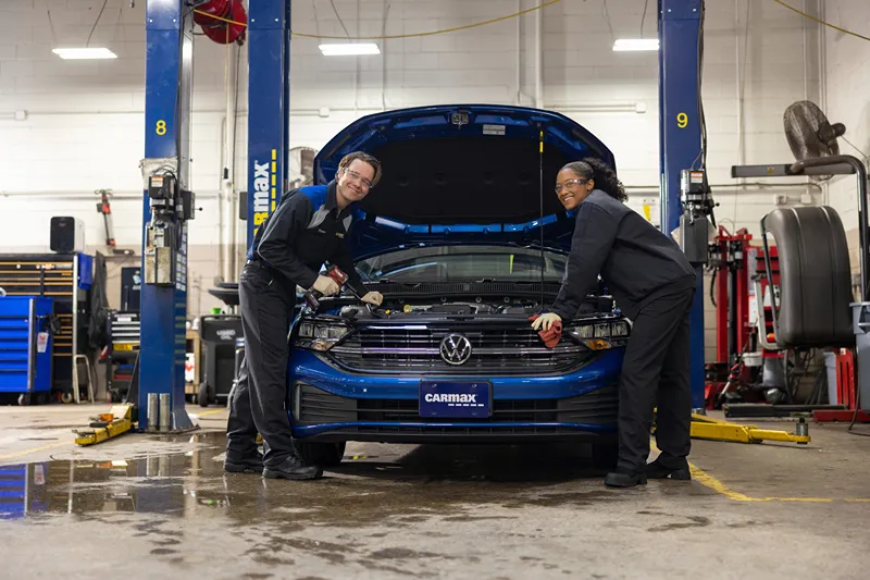 Two smiling CarMax auto technicians are working on a blue car in a service bay.