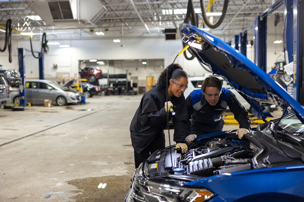 Two smiling auto technicians are inspecting a car engine with a flashlight in a service bay.