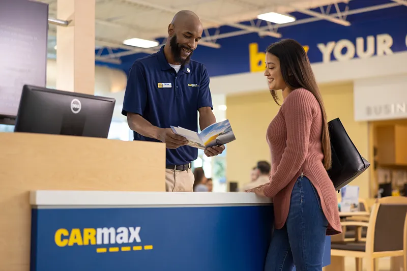 A CarMax sales consultant is smiling while assisting a customer at a service desk.
