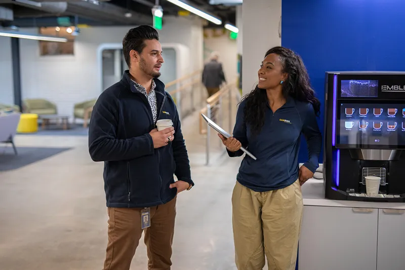 Two smiling CarMax corporate employees are chatting by a coffee machine in a modern office.