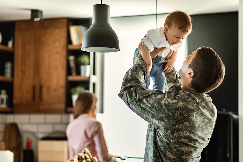 A father in military uniform is happily lifting his baby son in a bright kitchen.