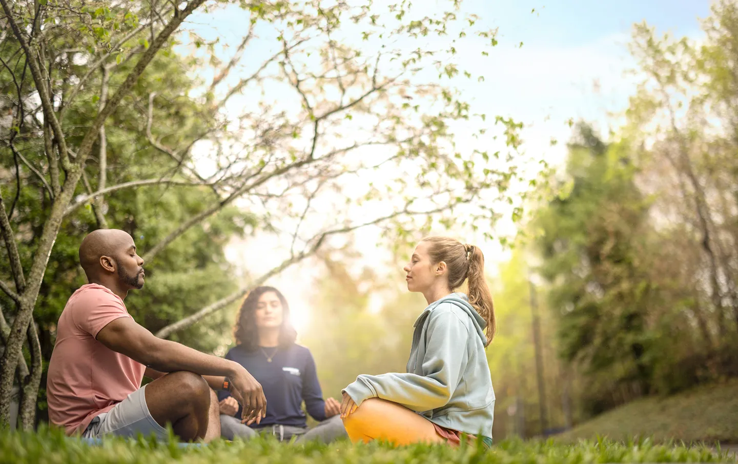 Three people are practicing mindfulness and meditation outdoors in a sunny park.