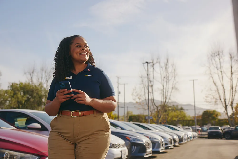 Smiling CarMax employee meeting with a customer in a bright office, discussing paperwork at a desk.