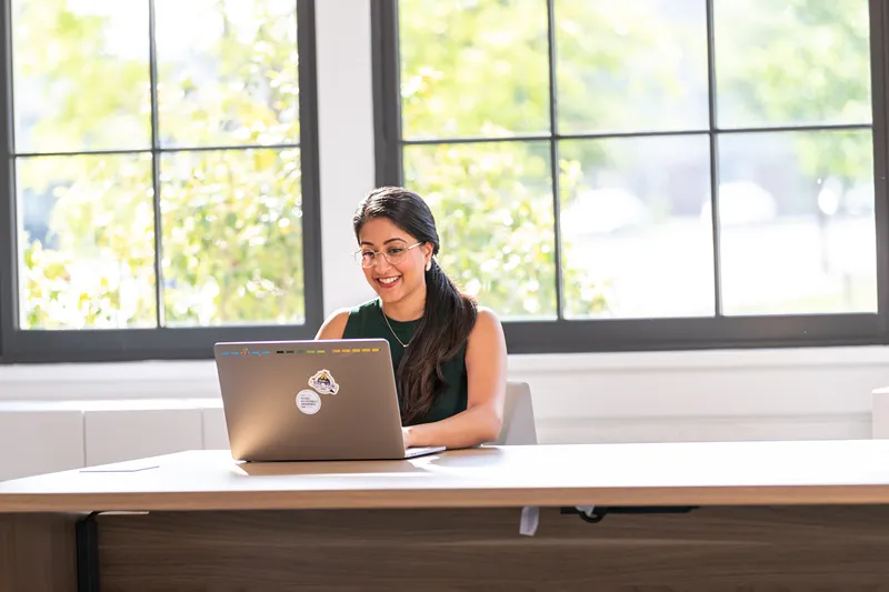 A smiling female intern or student is happily working on her laptop in a bright office space.