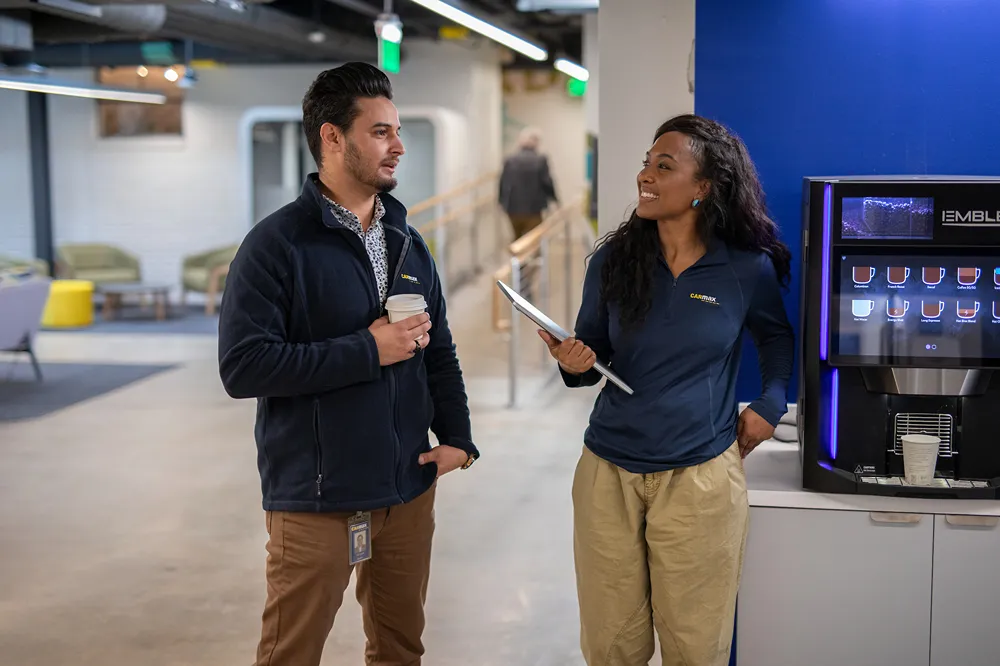 Two smiling CarMax corporate employees are chatting by a coffee machine in a modern office