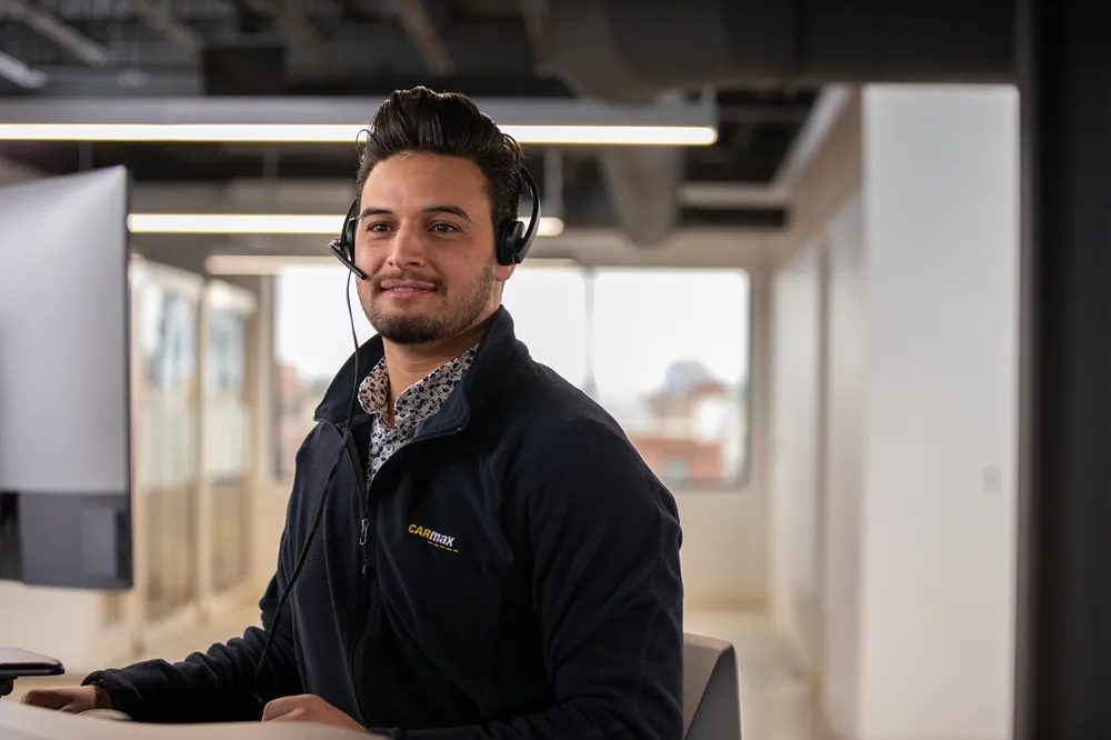 A smiling CarMax sales consultant is wearing a headset while working at his desk.