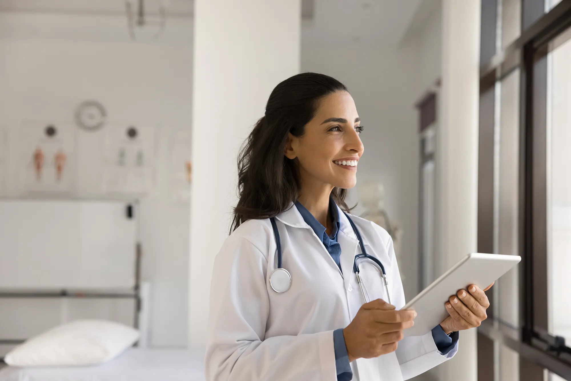 A doctory wearing a white coat and stethoscope looks onwards smiling