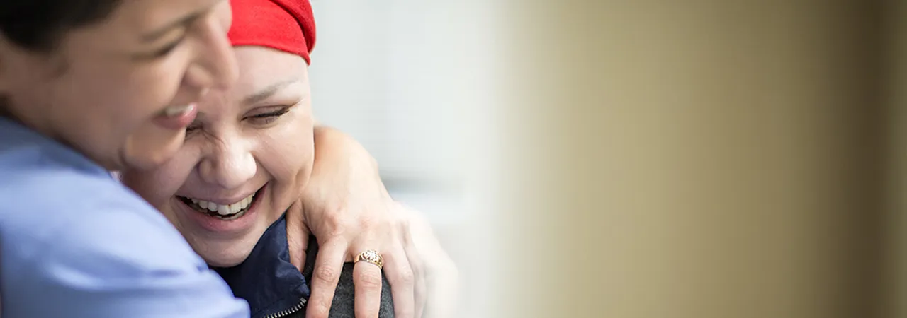 Nurse hugging and comforting a cancer patient wearing a red headscarf, both smiling warmly in a hospital setting.