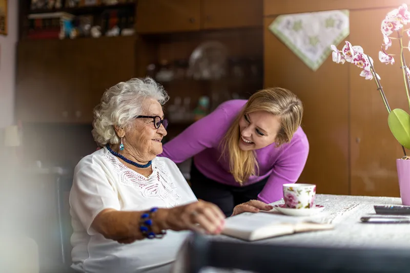 An elderly woman sitting in a chair at home, smiling warmly at a female healthcare professional who is kneeling beside them