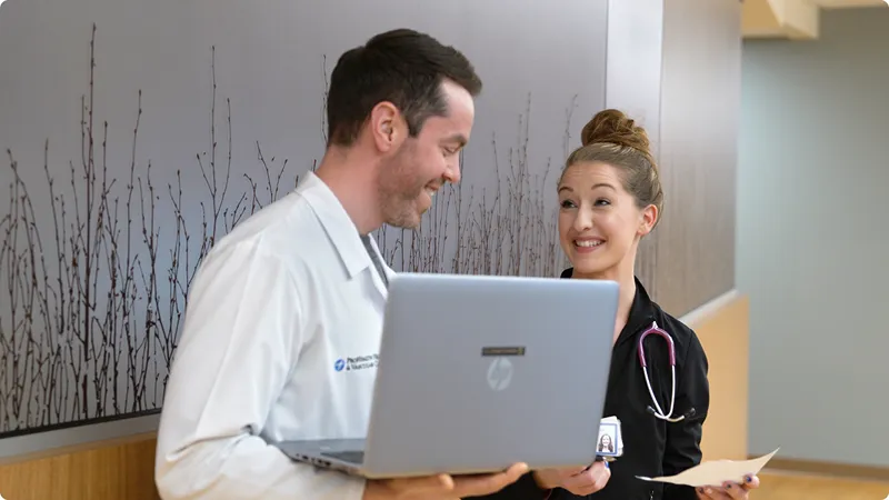Smiling medical resident in blue scrubs with a stethoscope and clipboard discusses a case with a medical student or junior resident