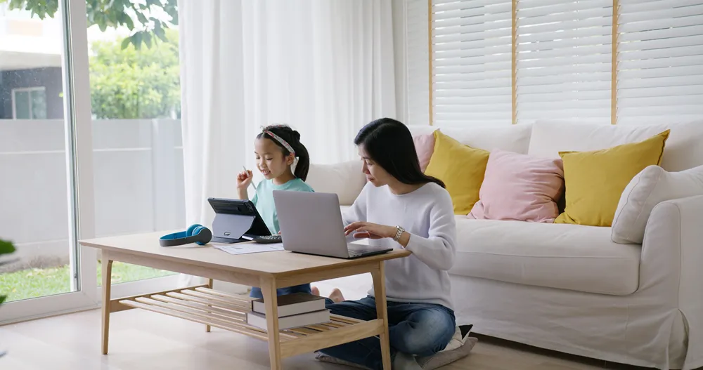 Mother working on laptop while child does homework at coffee table