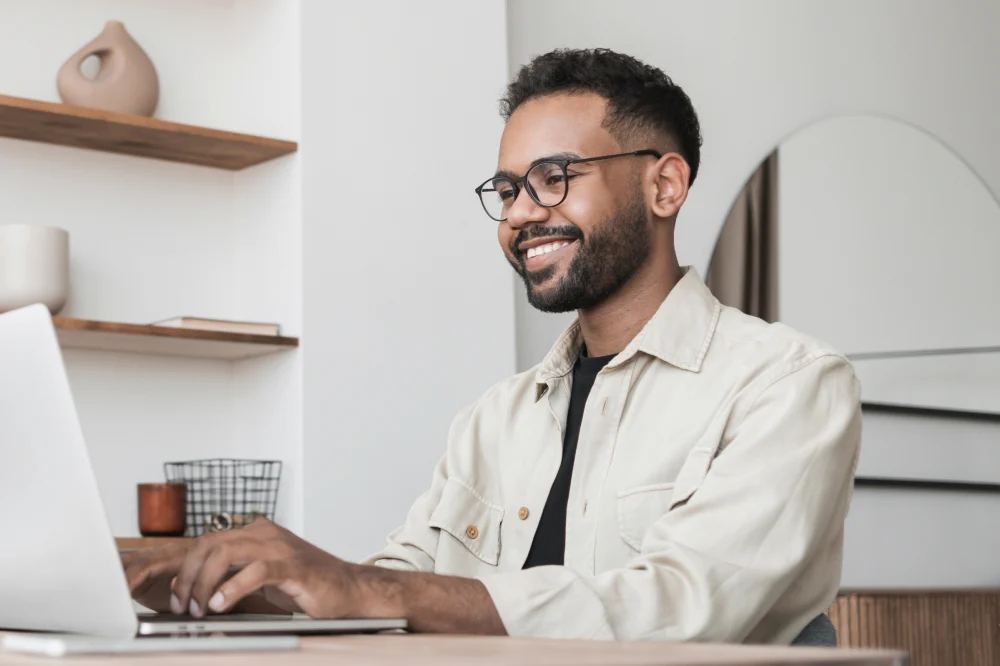 A young professional smiling while working remotely on a laptop in a modern home office