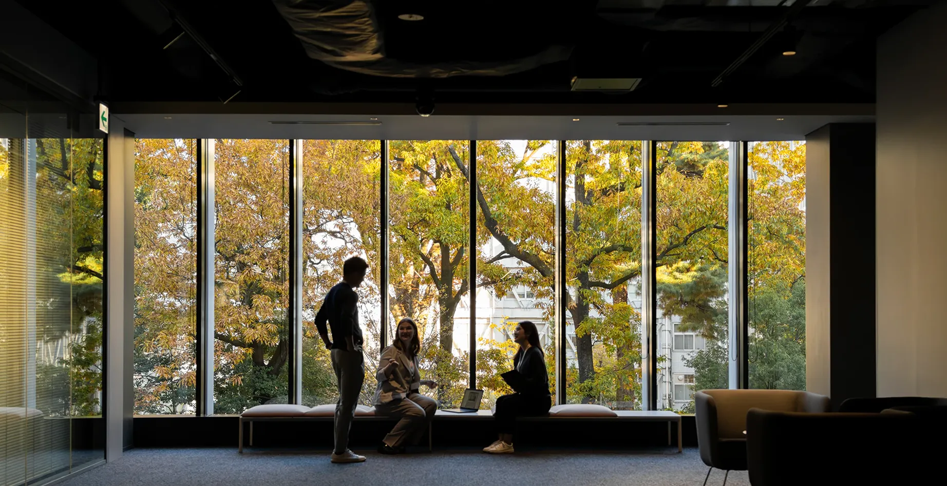 Colleagues having a casual conversation in modern office lounge with autumn trees visible through large windows