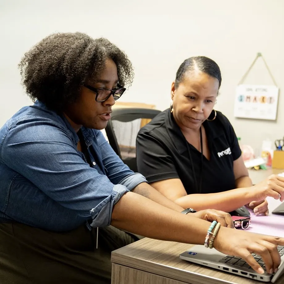 Two women collaborating at a desk with laptops, discussing work with focus and engagement.