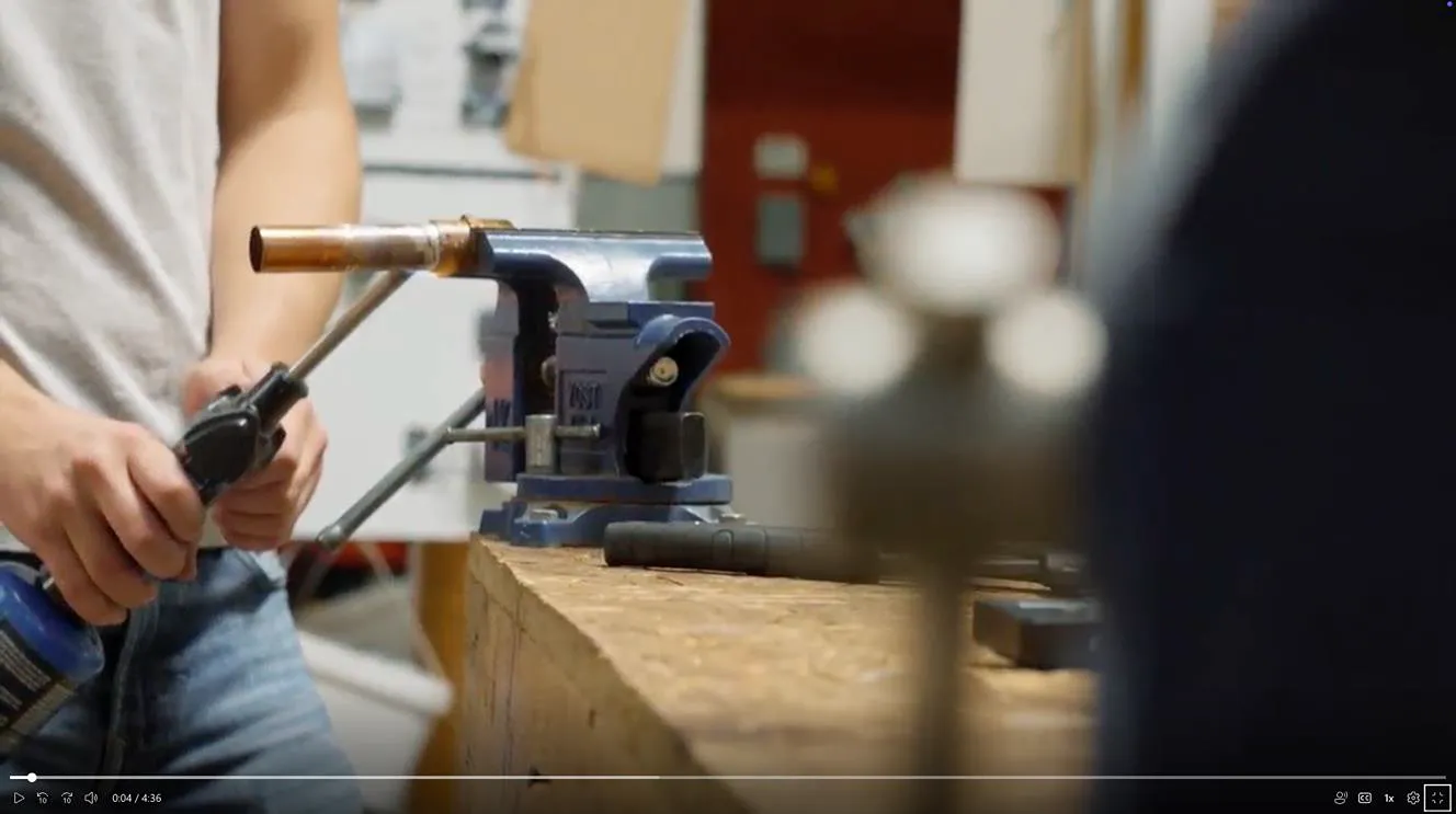 A person uses a torch to heat a copper pipe held in a vise on a workbench.