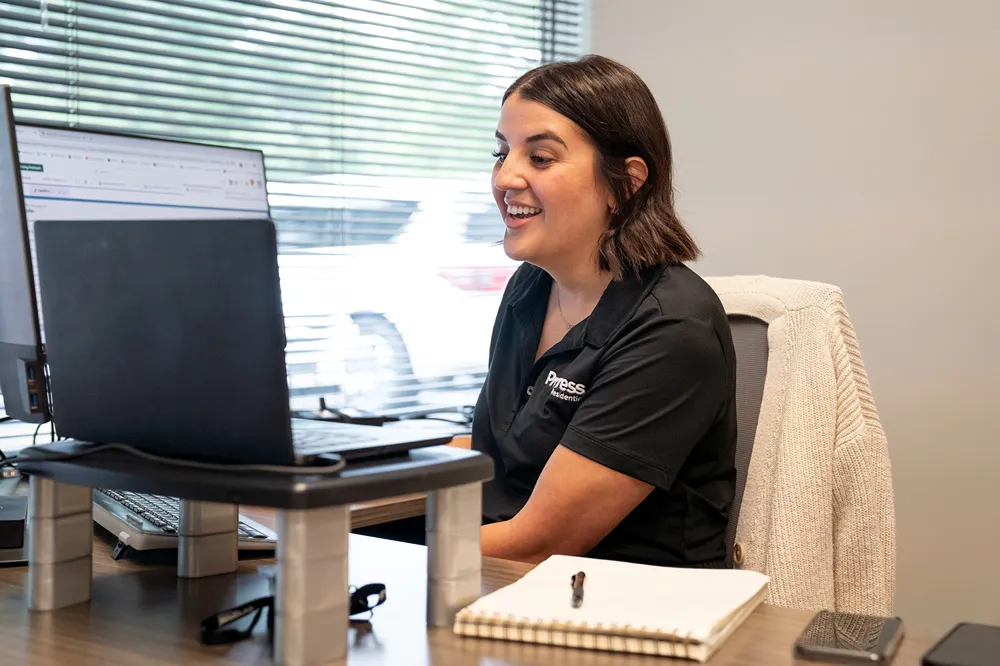 A smiling woman in a black polo shirt with a logo works at her desk with a laptop and computer monitor.