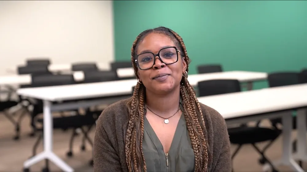 A young woman with braided hair and glasses smiles in front of a classroom setting