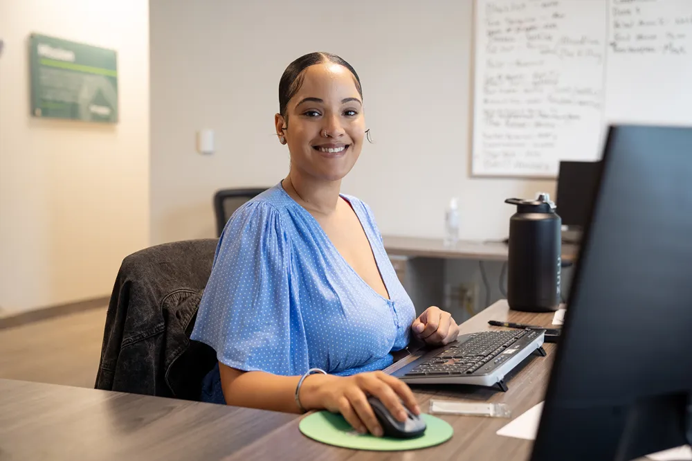 Young woman in a blue polka dot dress smiles at the camera while working at a computer.