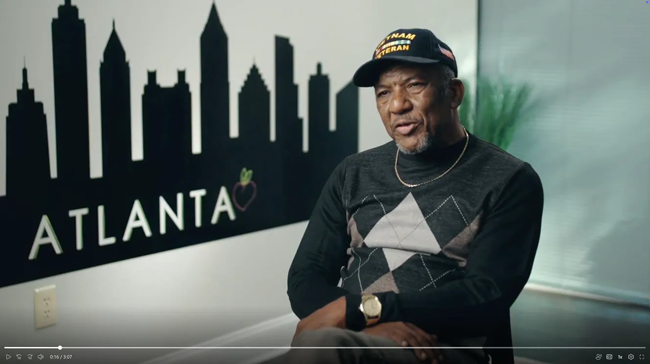 Older Black man wearing a Vietnam Veteran hat and argyle sweater sits in front of an Atlanta skyline wall graphic.